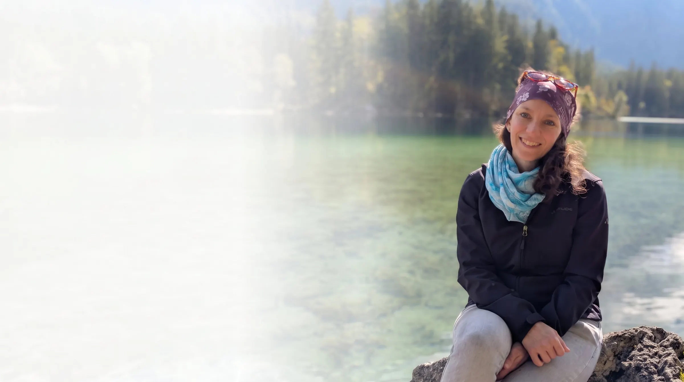 Maria Adamer sitzt lächelnd auf einem Stein an einem klaren Bergsee